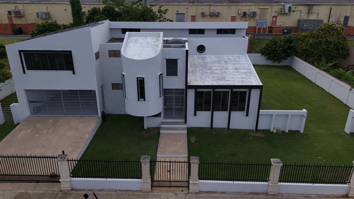 Aerial real estate photo of a modern white two-story home with a curved tower facade, gated entry and landscaped lawn in Puerto Rico
